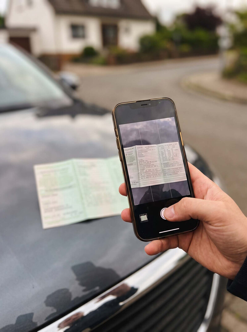 Fahrzeugschein auf einer Motorhaube, darüber ein Handy, das ihn fotografiert