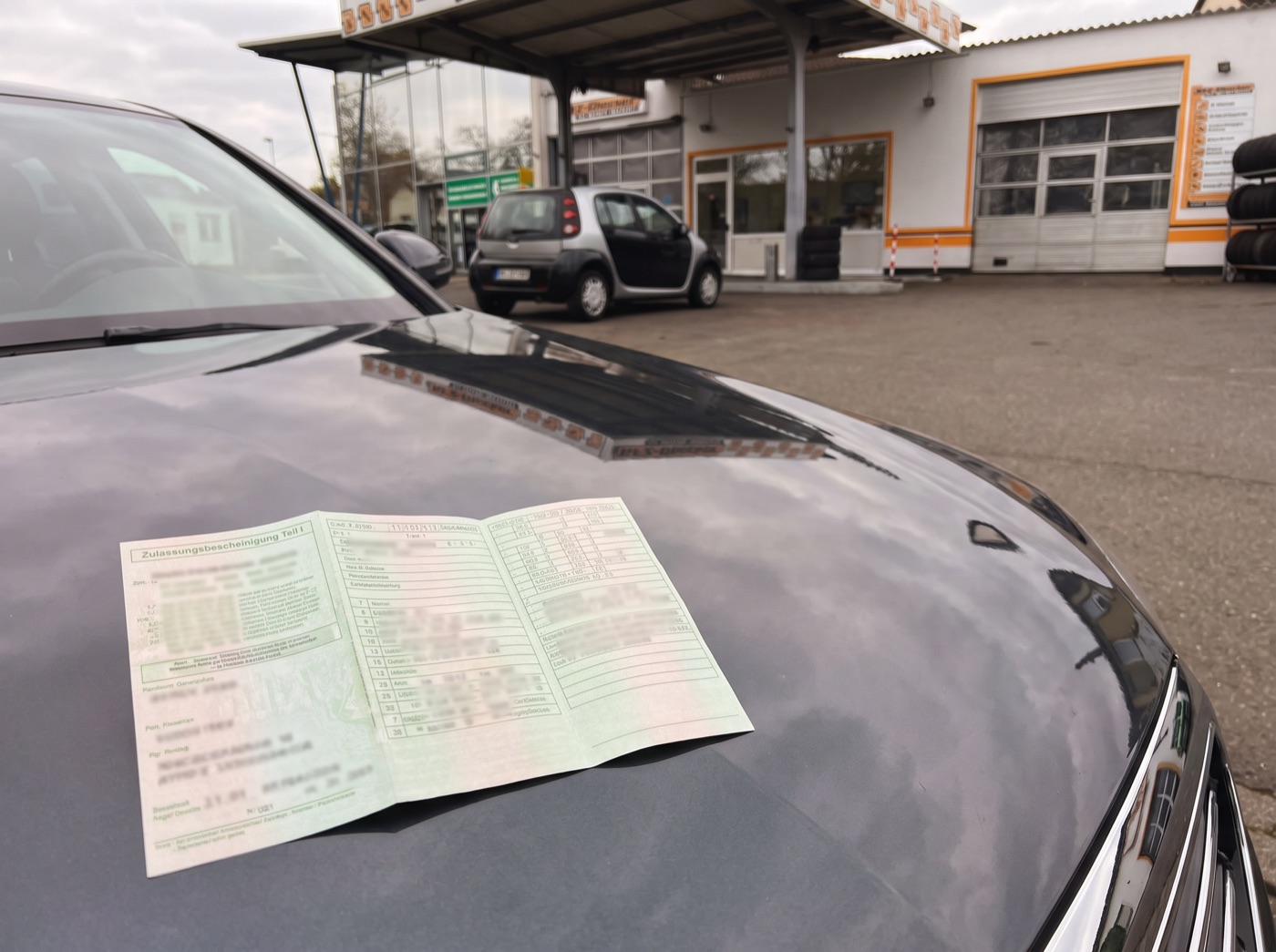 Aufgeklappter Fahrzeugschein auf einer Motorhaube, im Hintergrund eine Werkstatt